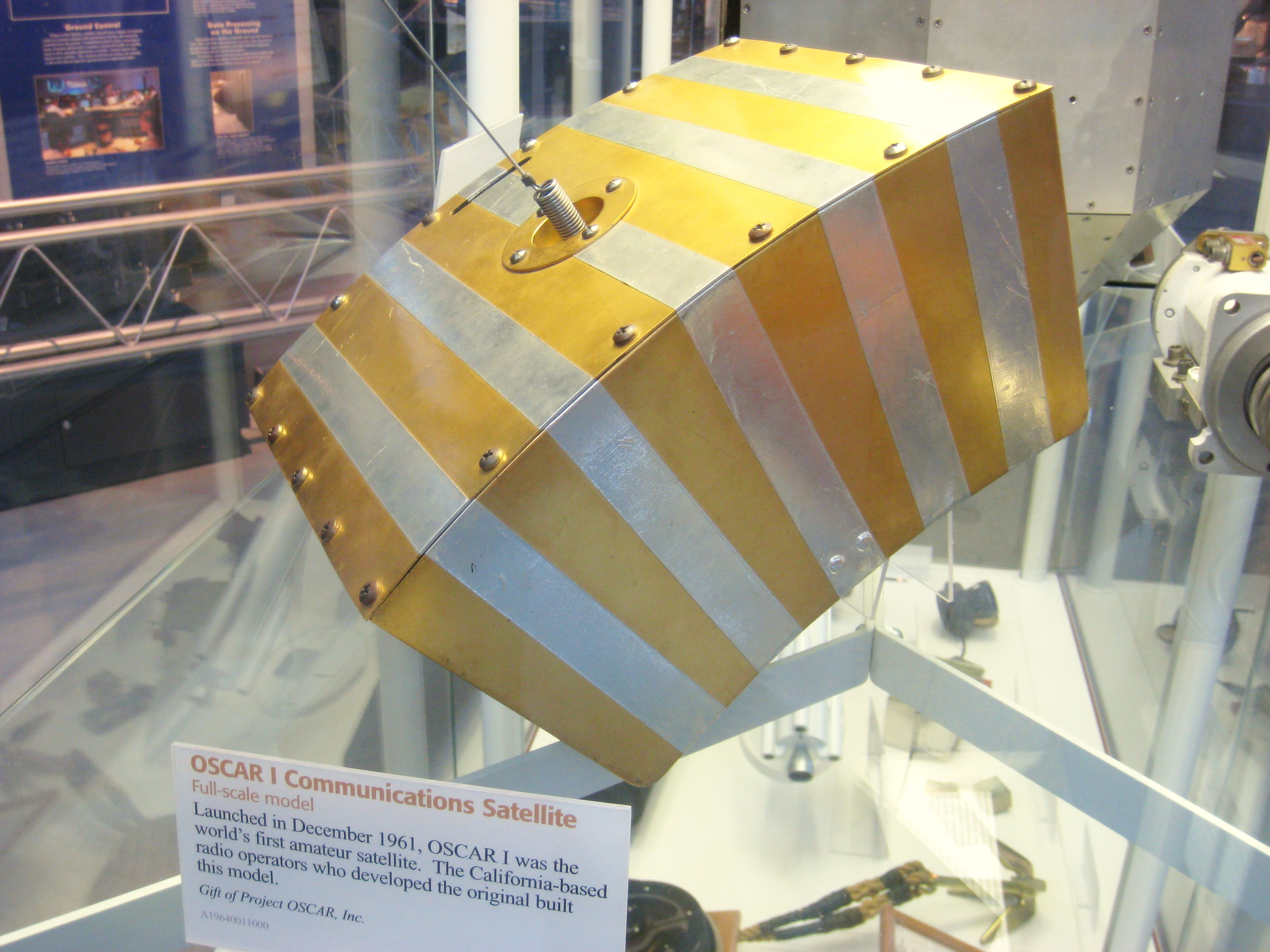 1) Kurzbeschreibung: Satellitenkörper mit kurzer Stabantenne in einer Glasvitrine.
<ol start=