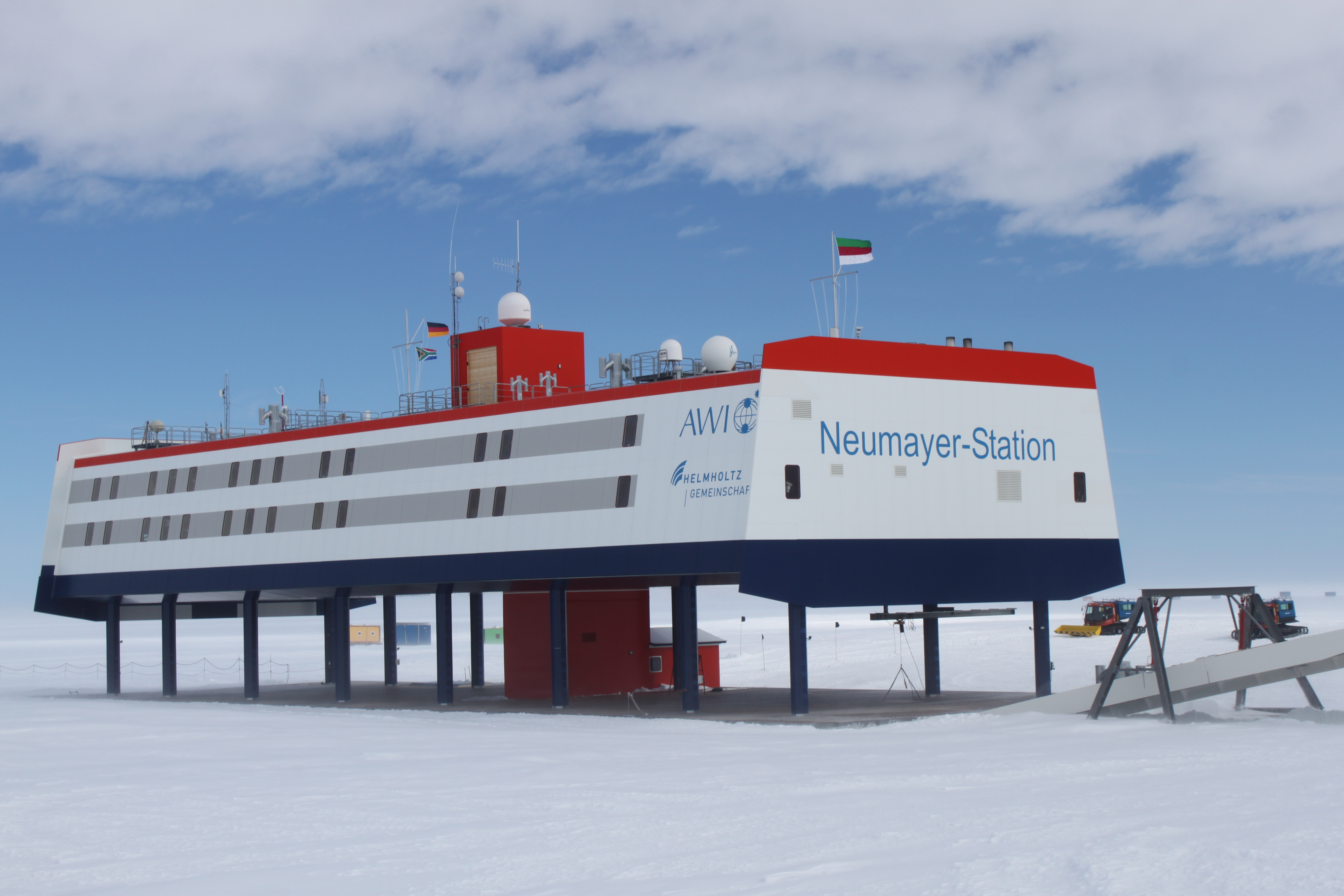 1) Kurzbeschreibung: Neumayer-Station in der Antarktis; langes, auf Stützen stehendes, weiß und grau gestreiftes Stationsgebäude mit rot abgesetzter Dachpartie und mit der Aufschrift „Neumayer-Station“ an der Stirnseite; auf dem Dach mehrere Masten, Antennen und Radome.
<ol start=