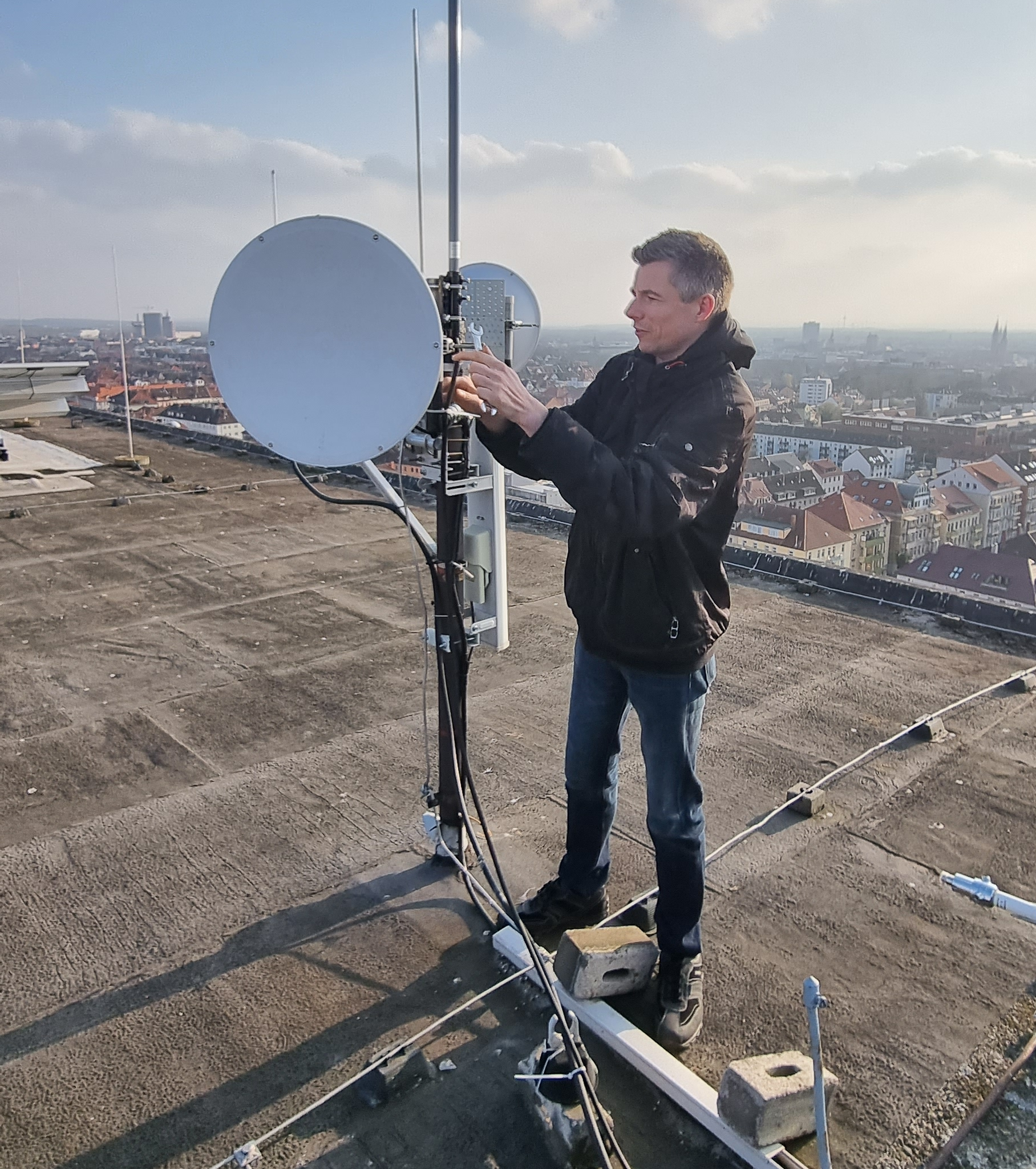 1) Kurzbeschreibung: Person auf einem Flachdach an einer Antennenanlage mit zwei Parabolspiegeln und Stabantennen.
<ol start=