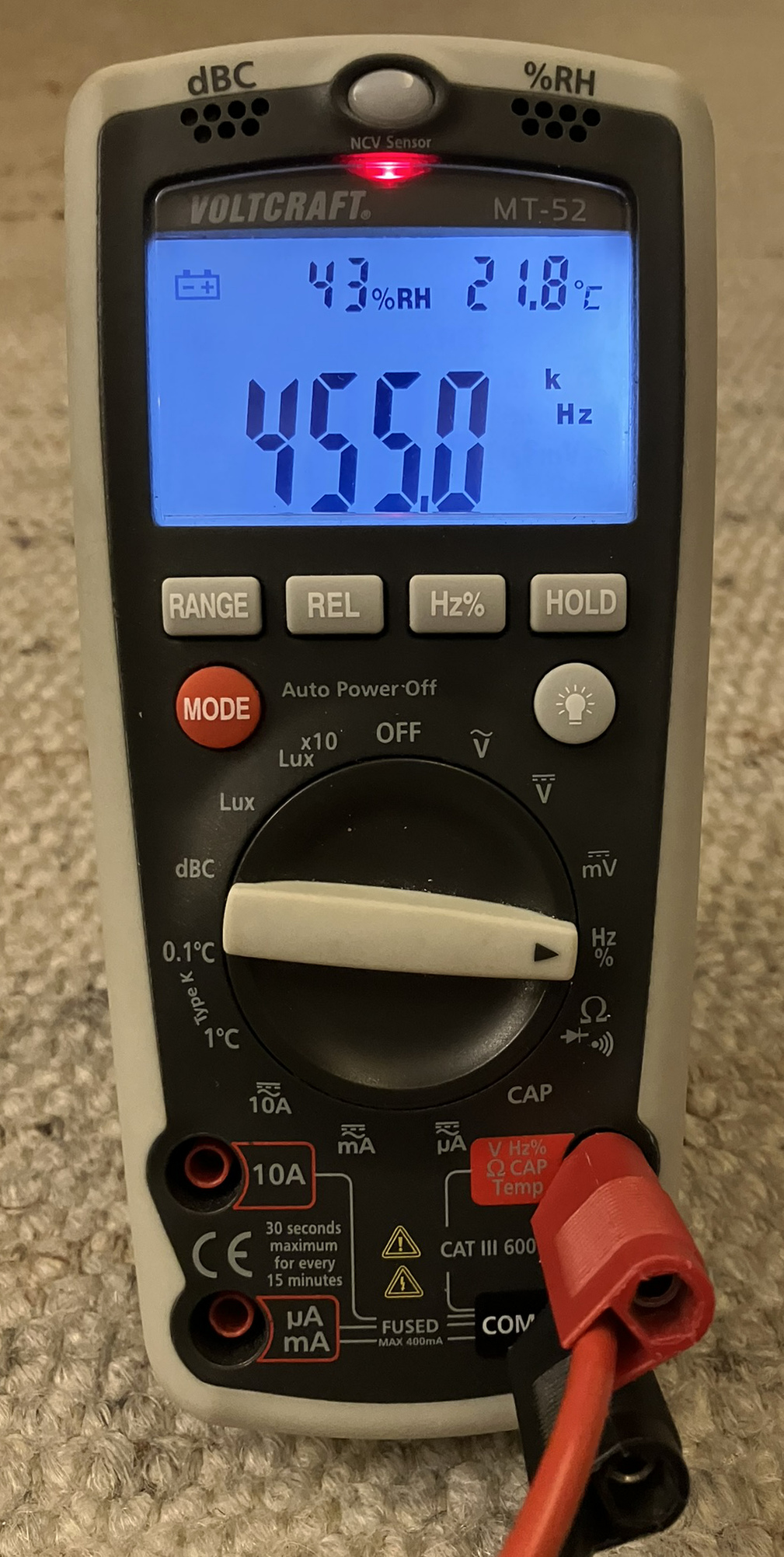 Dieser Alt-Text wurde noch nicht überprüft.
<p>Kurzfassung: Ein tragbares Digitalmultimeter mit blau beleuchtetem Display zeigt 455.0 kHz sowie 43 % relative Luftfeuchte und 21.8 °C an.</p>
<p>Detaillierte Beschreibung: Das Gerät im grauschwarzen Gummischutzgehäuse liegt auf einem beigen Teppich. Oben leuchtet eine rote LED neben der Aufschrift „NCV Sensor“; darüber sind die Hinweise „dBC“ und „%RH“ mit kleinen Lüftungsöffnungen zu sehen. Auf dem Display steht links ein Batteriesymbol, daneben „43%RH“ und rechts „21.8°C“; die großen Ziffern in der Mitte zeigen „455.0“, flankiert von den Einheiten „k“ und „Hz“. Unter dem Display befinden sich Tasten mit den Beschriftungen „RANGE“, „REL“, „Hz%“ und „HOLD“, links eine rote „MODE“-Taste und rechts eine Taste mit Lampensymbol. Der zentrale Drehschalter steht auf der rechten Seite in der Position mit der Beschriftung „Hz %“; weitere Skalen rundherum sind u. a. mit „V“, „mV“, „CAP“ und „Lux“ markiert. Unten sind vier Buchsen zu sehen: „10A“, „µA mA“, „COM“ und „V Hz Ω CAP Temp“; in „COM“ steckt ein schwarzer Stecker, in der rechten Buchse ein roter Stecker mit Kabel, daneben Sicherheits- und Messkategorienhinweise wie „CAT III 600V“ und „FUSED MAX 400mA“. Auf der Front sind außerdem die Markierungen „VOLTCRAFT“ und „MT-52“ sowie „Auto Power Off“ sichtbar.">
<figcaption>Abbildung E-10.9.1: Multimeter, das im Frequenzmessbereich $455 kHz$ anzeigt. Darüber erscheinen ein Symbol für niedrige Batteriespannung, die Luftfeuchtigkeit und die Temperatur. Diese Werte haben nichts mit der Frequenzmessung zu tun.</figcaption></p>
</figure>
</div>
</section>
<section></section>
<section><ul>
<li>Anzeige der Frequenz</li>
<li>Bei älteren Geräten steht hinten ein $10^x$-Multiplikator
</left></li>
</ul>
<div class=