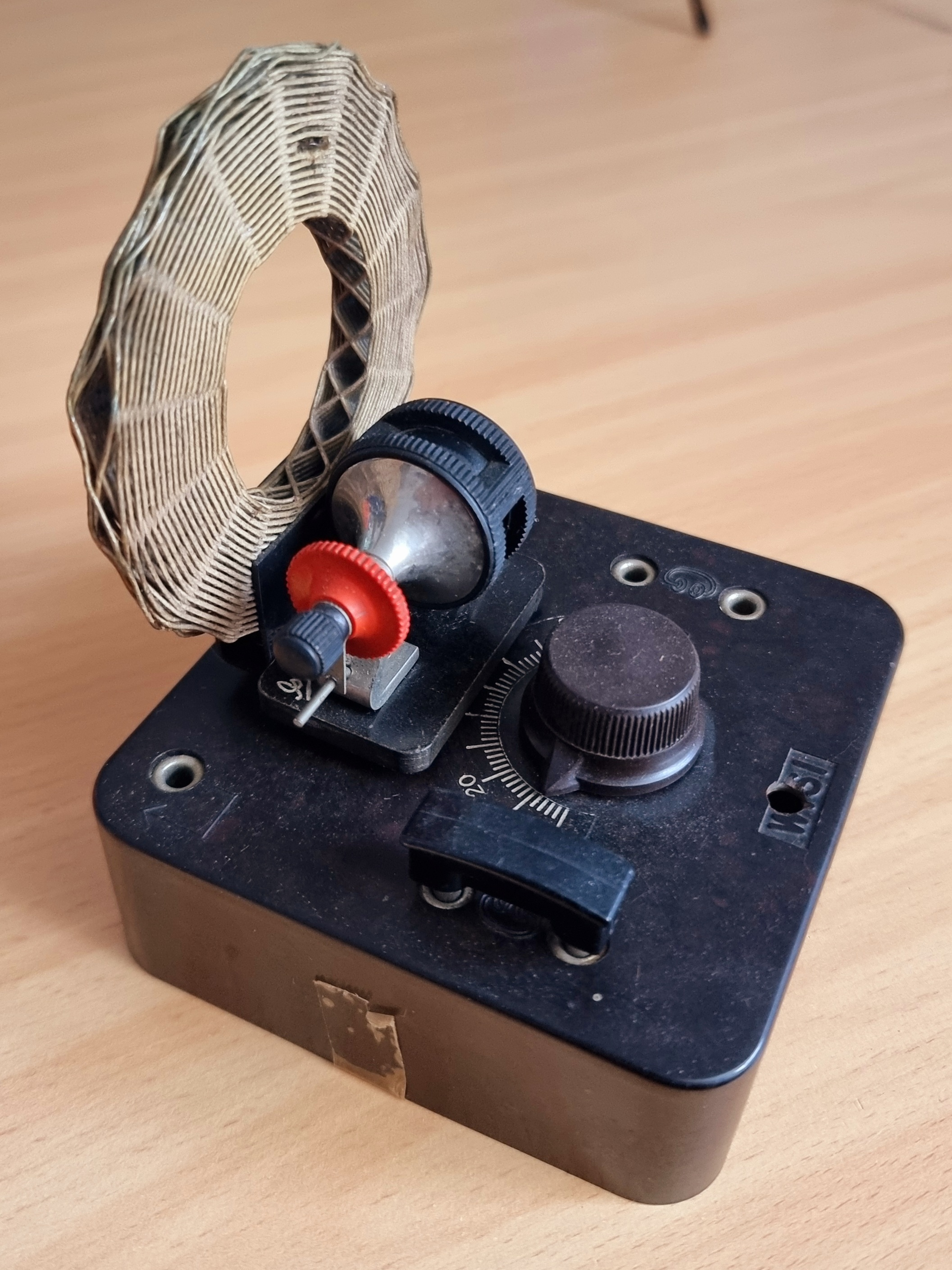 Dieser Alt-Text wurde noch nicht überprüft.
<p>Kurzfassung: Ein schwarzes, älter wirkendes Tischgerät mit senkrechter Ringspule und mehreren Einstellelementen steht auf einer hellen Holzoberfläche.</p>
<p>Detaillierte Beschreibung: Auf einem rechteckigen, dunkelbraunen/ schwarzen Gehäuse (mit abgenutzter Oberfläche) sitzt links eine flache, aufrecht stehende Ringspule aus eng gewickeltem, hellem Drahtband mit rautenförmigem Muster. Davor befinden sich ein kleiner schwarzer Einstellmechanismus mit rotem gerändelten Rädchen und ein glänzendes, halbkugeliges Metallteil. Rechts ist ein großer, gerändelter Drehknopf neben einer halbkreisförmigen Skala mit Markierungen (u. a. 0–20), darüber ein schwarzer Schieber als Zeiger. In der rechten oberen Gerätefläche liegen zwei versenkte Metallschrauben/Anschlüsse; an zwei Ecken sind Bohrungen sichtbar. An der Frontseite klebt ein kleines Stück vergilbtes Klebeband. Der Hintergrund ist unscharf, die Holzplatte zeigt feine Maserung.">
<figcaption>Abbildung NEA-13.2.1: Detektor-Empfänger</figcaption></p>
</figure>
</aside>
Beim Detektor-Empfänger wird ein Parallel-Schwingkreis, bestehend aus Spule und abstimmbarem Kondensator, durch ein empfangenes Rundfunksignal <a class=