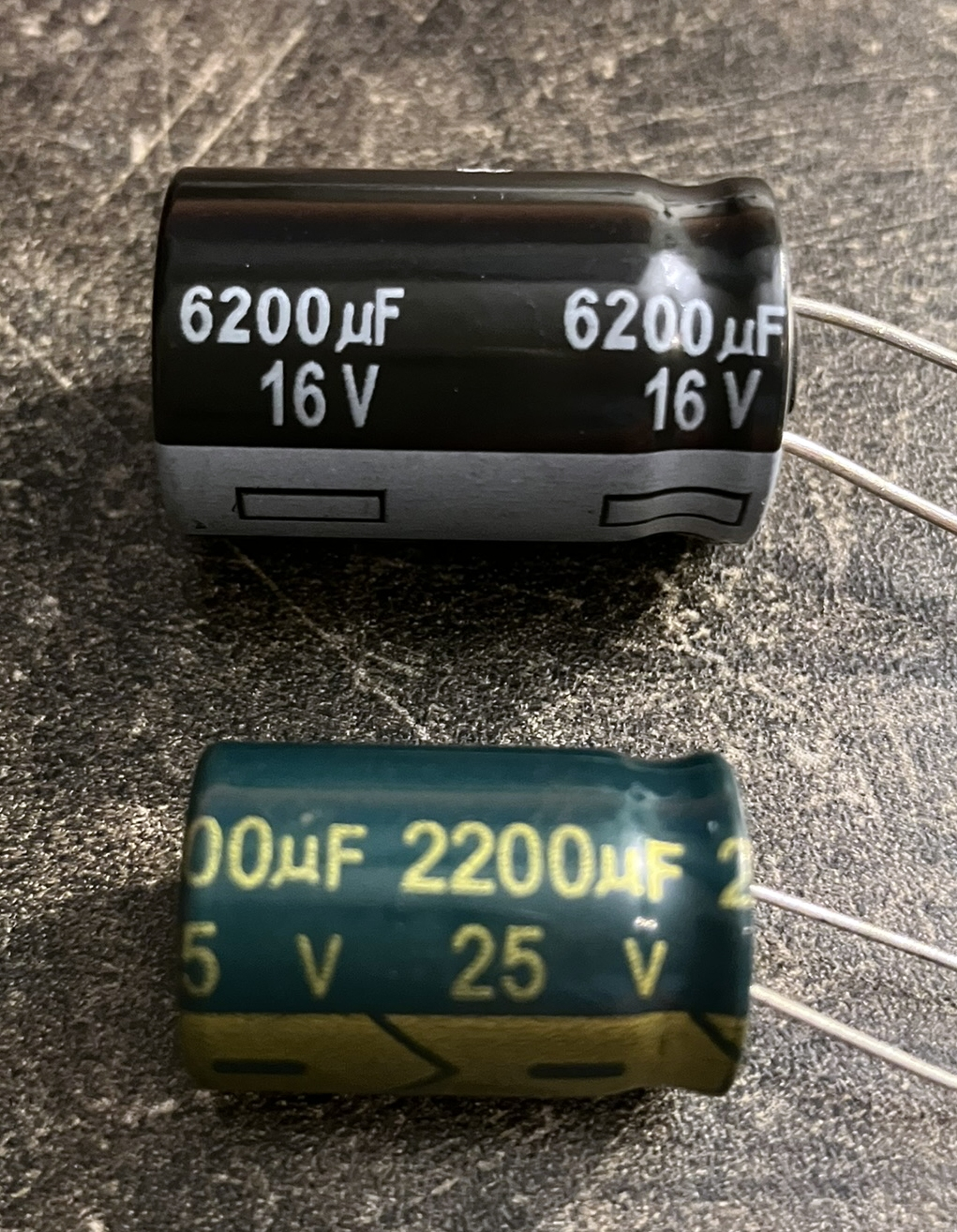 Dieser Alt-Text wurde noch nicht überprüft.
<p>Kurzfassung: Zwei liegende Elektrolytkondensatoren mit Drahtanschlüssen auf einer dunklen, rauen Arbeitsfläche.</p>
<p>Detailbeschreibung: Oben ein größerer, zylindrischer Kondensator mit schwarzem Gehäuse und grauem Ring am unteren Ende; in weißer Schrift steht mehrfach „6200 µF 16 V“, die beiden Anschlussdrähte ragen nach rechts heraus. Darunter ein etwas kleinerer, dunkelgrüner Kondensator mit gelb-goldener Aufschrift „2200 µF 25 V“ und zusätzlichen Markierungen auf einem olivfarbenen Streifen; auch hier führen zwei Drähte nach rechts. Die Oberfläche zeigt Kratzer und Staub, das Licht erzeugt glänzende Reflexe auf den Kondensatoren, die nahezu parallel zueinander liegen.">
<figcaption>Abbildung E-3.3.2: Elektrolytkondensatoren mit den Spannungsfestigkeiten 16 Volt und 25 Volt</figcaption></p>
</figure>
</aside>
Eine weitere Kenngröße ist der *Spitze-Spitze-Wert*. Das ist der Unterschied zwischen dem höchsten und dem niedrigsten Ausschlag. Für Sinusförmige Wechselspannungen gilt:
<p>$U_\text{SS} = 2\cdot \hat{U}$.</p>
<section class=