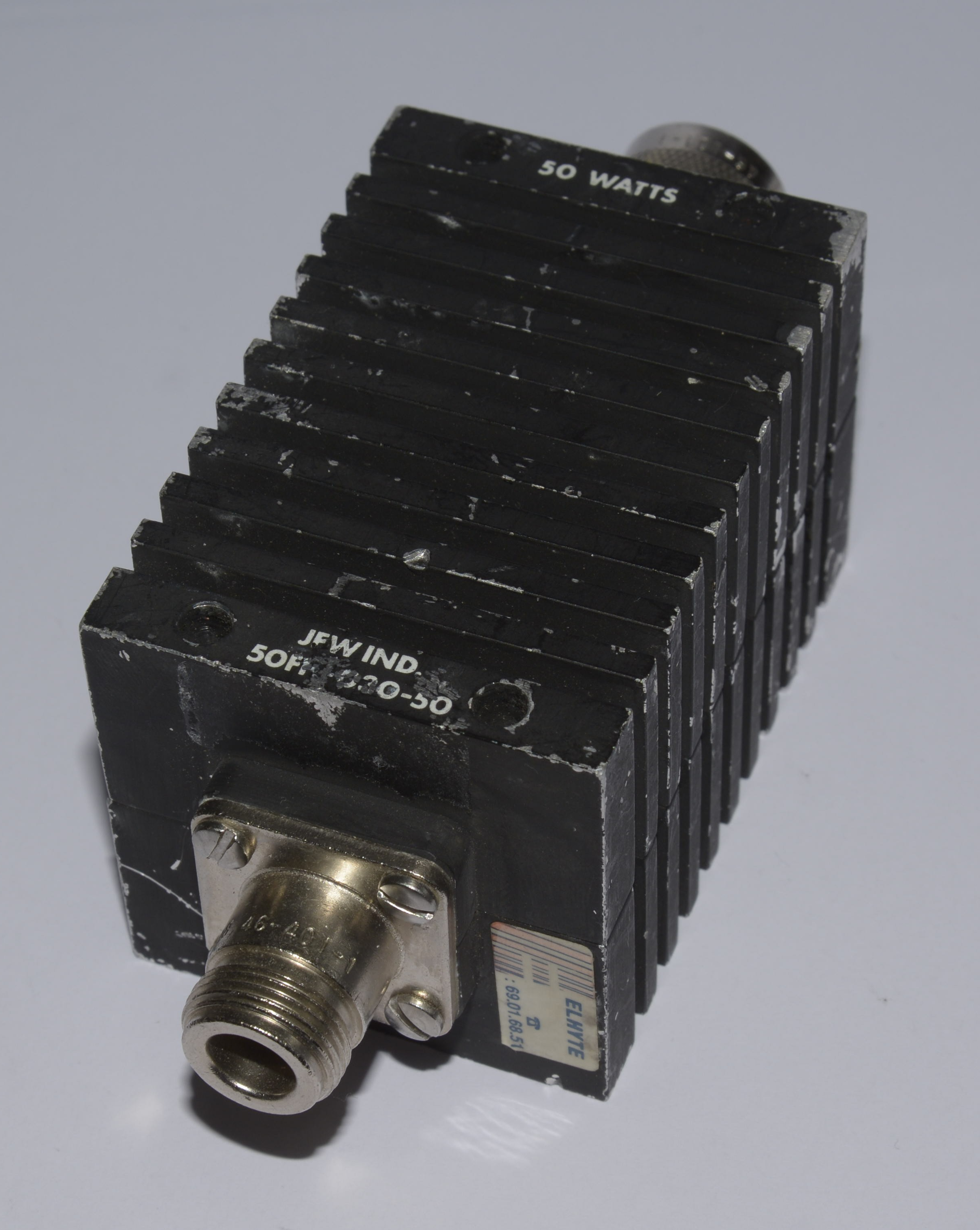 Dieser Alt-Text wurde noch nicht überprüft.
<p>Kurzfassung: Schwarzer, quaderförmiger Metallblock mit dichten Kühlrippen, der Aufschrift „50 WATTS“ und Koaxialanschlüssen an beiden Enden.</p>
<p>Detaillierte Beschreibung: Das Objekt liegt schräg auf einem hellgrauen Hintergrund und besteht aus einem schwarzen, gerippten Kühlkörper mit mehreren parallelen Längsrippen. An der Vorderseite sitzt ein koaxialer Einbausteckverbinder auf einer rechteckigen Metallflanschplatte, befestigt mit zwei Schrauben; die Lackierung ist an Kanten und Flächen sichtbar angeschlagen. Auf der Stirnseite sind die Worte „JFW IND.“ sowie eine teils abgeriebene Modellbezeichnung zu lesen. Auf der Oberseite steht „50 WATTS“. Auf der rechten Seitenfläche klebt ein kleiner Aufkleber mit der Aufschrift „ELMITE“ und einer Zahlenfolge. Hinten ist ein weiterer koaxialer Anschluss teilweise sichtbar. Das Licht fällt von oben links ein und erzeugt Schatten in den Rippenzwischenräumen.">
<figcaption>Abbildung NEA-13.14.1: Ein Dämpfungsglied</figcaption></p>
</figure>
<figure class=