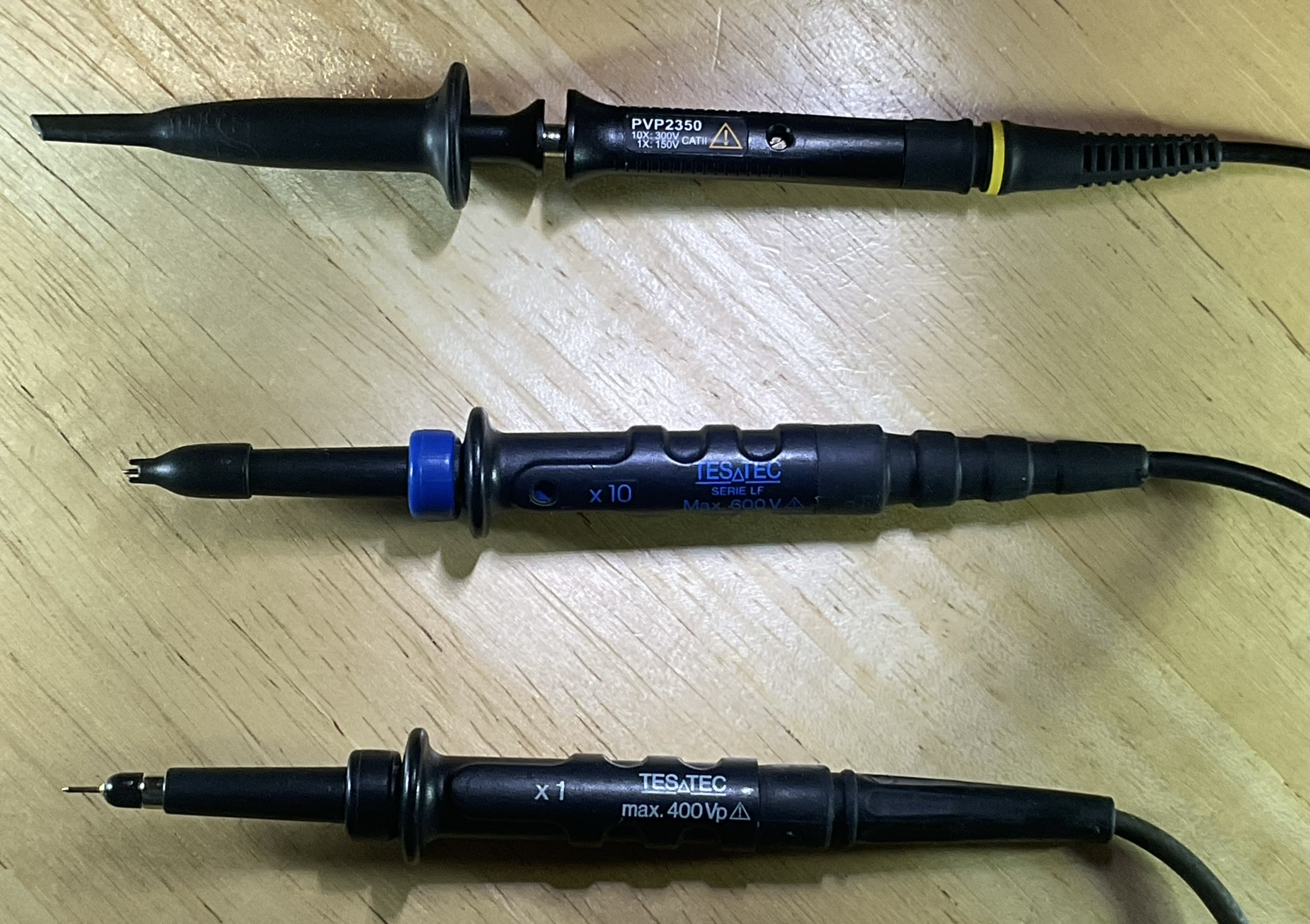 Dieser Alt-Text wurde noch nicht überprüft.
<ol>
<li>
<p>Kurzzusammenfassung: Drei schwarze Oszilloskop‑Tastköpfe liegen parallel auf einer hellen Holzoberfläche.</p>
</li>
<li>
<p>Detailbeschreibung: Die drei Sonden sind horizontal ausgerichtet, mit den Spitzen nach links und den Kabeln nach rechts. Oben befindet sich ein schwarzer Tastkopf mit der Aufschrift „PVP2350“, Warnsymbol und gelbem Ring an der Zugentlastung; die Spitze ist mit einer länglichen schwarzen Schutzkappe versehen. In der Mitte liegt ein schwarzer Tastkopf mit blauem Einstellring und der blauen Beschriftung „x10“, „TESATEC“, „SERIE LF“, „Max. 600V“; an der Spitze ist eine kleine Haken- oder Steckspitze sichtbar, teils von einer kurzen Kappe umgeben. Unten liegt ein weiterer schwarzer Tastkopf mit weiß/blauer Beschriftung „x1“, „TESATEC“, „max. 400Vp“; hier ist eine feine, nadelförmige Metallspitze freiliegt. Die Gehäuse haben gerippte Griffflächen, die Kabel führen nach rechts aus dem Bild, und die Holzoberfläche zeigt deutliche Maserung."></p>
 <figcaption>Abbildung NEA-6.15.4: Tastköpfe mit verschiedenen Prüfspitzen. Die Krokodilklemmen wurden für diese Aufnahme abgenommen.</figcaption>
</li>
</ol>
</figure>
</aside>
Die einfachsten Tastköpfe verbinden die Prüfspitze direkt mit dem Messeingang. Man spricht von 1:1-Tastköpfen, weil die an der Spitze anliegende Spannung unverändert zum Oszilloskop gelangt. Tastköpfe für hohe Frequenzen sind aufwändiger gebaut. Sie teilen die Eingangsspannung auf einen kleineren Wert, oft ein Zehntel, herunter. Wenn man mit so einem 10:1-Tastkopf eine Spannung von 10 Volt misst, wird auf dem Bildschirm 1 Volt angezeigt.
<aside class=
