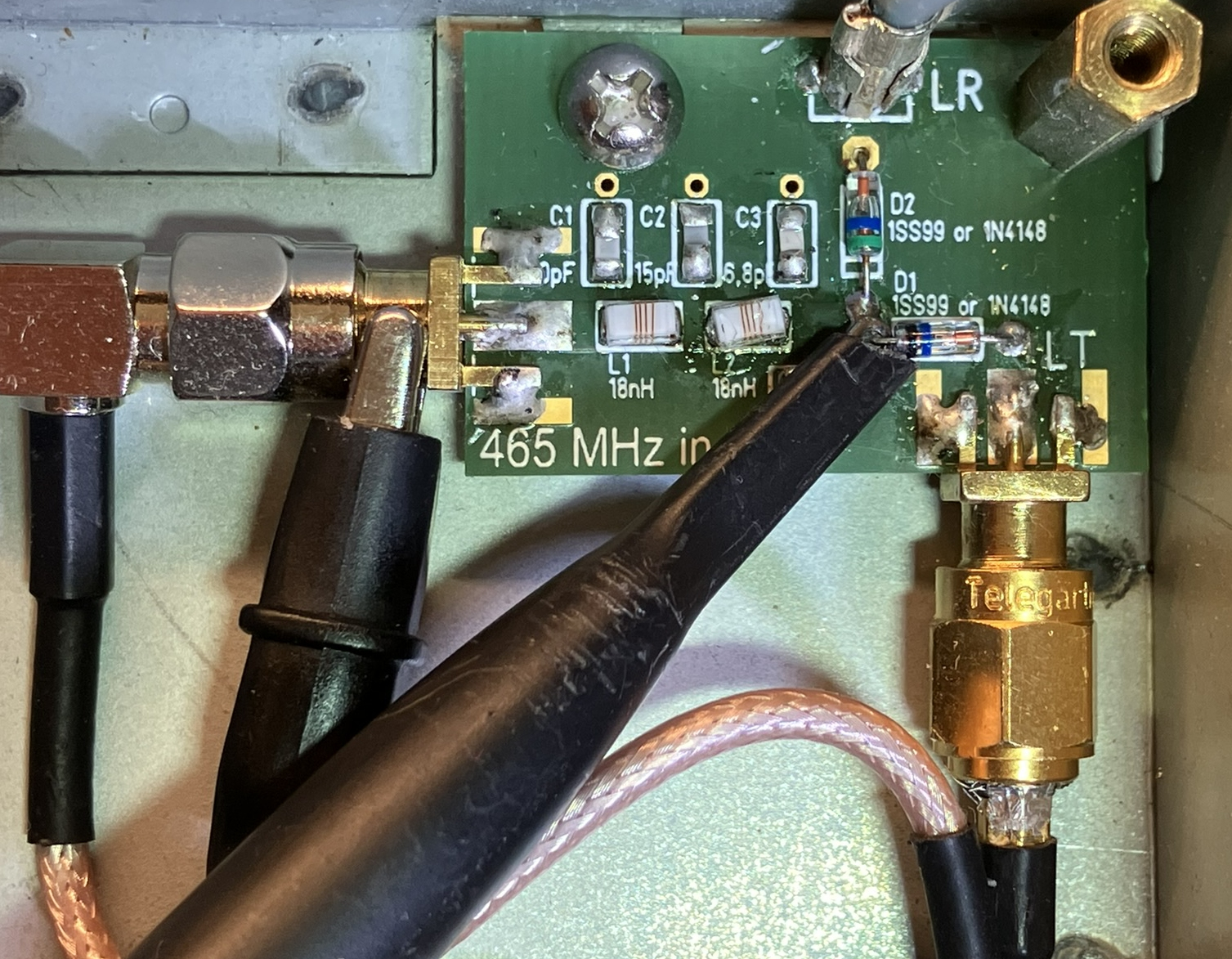 Dieser Alt-Text wurde noch nicht überprüft.
<ol>
<li>
<p>Kurzfassung: Geöffnete Elektronikbaugruppe in einem Metallgehäuse mit einer kleinen grünen Leiterplatte, mehreren Koax‑Steckverbindern und angesetzten Messleitungen.</p>
</li>
<li>
<p>Detaillierte Beschreibung: In der rechten Bildhälfte liegt eine grüne Leiterplatte mit weißer Beschriftung; sichtbar sind u. a. Bezeichnungen C1, C2, C3, L1, L2, D1, D2 sowie der Aufdruck „465 MHz in“. Neben den Markierungen für D1/D2 sind zwei kleine bedrahtete Bauteile mit Farbringen eingelötet; daneben liegen weitere bestückte und unbestückte SMD‑Plätze. Am rechten Rand ragt ein goldfarbener, verschraubter Koax‑Steckverbinder (SMA‑Bauform) nach unten aus dem Gehäuse; auf dem Sechskant steht „Telegärtner“. Links führt ein schwarzes Koaxkabel über einen rechtwinkligen metallischen Adapter auf die Platine. Über der Platine sind zwei schwarze Tastköpfe oder Leitungen angesetzt; ein spitzer schwarzer Prüfstift berührt einen Lötpunkt neben den beschrifteten Bauteilen. Die Platine ist mit mehreren Lötstellen, etwas Flussmittelrückständen und einer Schraube am Gehäuse befestigt; der Hintergrund besteht aus blankem Metall. Ein rosafarben ummanteltes Koaxkabel verläuft von unten rechts zum goldfarbenen Stecker."></p>
 <figcaption>Abbildung NEA-6.15.3: Messung mit einem Tastkopf. Zwischen den Dioden D1 und D2 ist die Prüfspitze zu sehen und weiter links die Krokodilklemme für die Masseverbindung.</figcaption>
</li>
</ol>
</figure>
</aside>
<aside class=