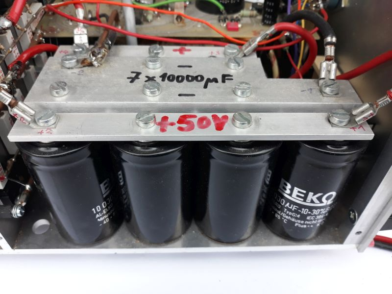 Dieser Alt-Text wurde noch nicht überprüft.
<ol>
<li>
<p>Kurze Zusammenfassung:
Vier große, schwarze Elektrolytkondensatoren sind in einem Metallgehäuse montiert und über zwei verschraubte Aluminiumschienen mit roten Anschlusskabeln verbunden, mit handschriftlichen Plus/Minus-Markierungen und der Aufschrift „+50V“ sowie „7 × 10000 µF“.</p>
</li>
<li>
<p>Detaillierte Beschreibung:
Im Vordergrund befinden sich vier zylinderförmige, schwarze Kondensatoren in einer Reihe, mit hellen Aufdrucken wie „10000 µF -10…+30%“ und dem Markenaufdruck „BEKO“; sie stehen in einer Metallwanne. Darüber liegen zwei parallel verlaufende, silberfarbene Aluminiumschienen, die mit mehreren Sechskantschrauben und Unterlegscheiben verschraubt sind. Auf der oberen Schiene sind handschriftliche Kennzeichnungen zu sehen: ein rotes „+“, ein schwarzes „-“, sowie in schwarzer Schrift „7 × 10000 µF“. Auf der unteren Schiene steht in roter Schrift „+50V“. An den Enden und an mehreren Punkten der Schienen sind rote Leitungen mit Ringkabelschuhen angeschlossen; einige Anschlüsse sind mit transparenten Isolierschläuchen versehen. Im Hintergrund sind weitere Bauteile, Leiter, Lötstellen und mehrfarbige Kabel innerhalb des Metallchassis sichtbar. Die Szene zeigt eine geordnete, verschraubte Verdrahtung, nummerierte Markierungen an einzelnen Schrauben und ein insgesamt sauber aufgebautes Modul innerhalb eines größeren elektronischen Geräts."></p>
 <figcaption>Abbildung NEA-9.7.2:  Parallelschaltung von $7 \times 10000&nbsp;µF$ in einem Endstufennetzteil</figcaption>
</li>
</ol>
</figure>
</aside>
Als Verständnistest kann die nächste Aufgabe verwendet werden.
<section class=