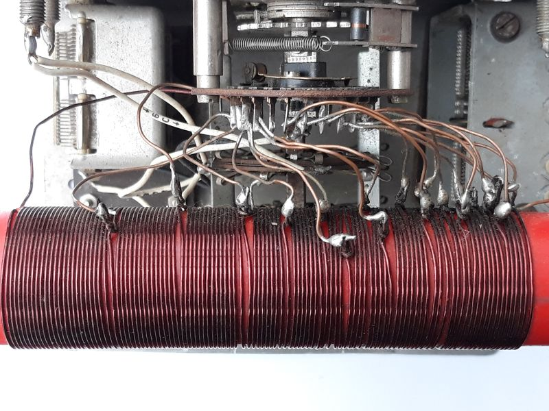 Dieser Alt-Text wurde noch nicht überprüft.
<p>Zusammenfassung: Nahaufnahme des Innenraums eines elektronischen Geräts mit einer dicht gewickelten roten Zylinderspule und zahlreichen Drahtabgriffen, die zu einem mehrstufigen Drehschalter im Metallchassis führen.</p>
<p>Detaillierte Beschreibung: Im unteren Bildbereich liegt horizontal eine rote, zylindrische Spule, eng mit dunkelrotem Kupferdraht bewickelt. Entlang der oberen Spulenseite sind viele Abgriffe sichtbar: dünne, bräunliche lackierte Drähte, die an einzelnen Windungen angelötet sind; an den Lötstellen sitzen silbrig glänzende Lottropfen, teils mit dunklen Verfärbungen an der Umgebung. Die Abgriffe verlaufen bogenförmig nach oben und vorne zu einem gestapelten, kreisförmigen Drehschalter mit mehreren Isolatorscheiben und radial angeordneten Lötfahnen. In der Schaltermitte sitzt eine Metallwelle mit Federn und kleinen mechanischen Teilen. Links und rechts erkennt man das graue Metallchassis mit Schrauben, Federn und Halterungen; einzelne weiße und graue Leitungen führen in den Aufbau. Der Hintergrund ist überwiegend grau-metallisch, unter der Spule ist ein heller, weißer Untergrund zu sehen. Der Gesamteindruck ist technisch, gebraucht und funktional, mit leicht unordentlich geführten Drähten.">
<figcaption>Abbildung EA-6.8.1: Spule mit 14 Anzapfungen in einem selbstgebauten Antennenanpassgerät</figcaption></p>
</figure>
Die Teilabschnitte zwischen den Anzapfungen können wie einzelne Spulen gesehen werden, deren Induktivität, durch Umschaltung der Anzapfungen,  in Schritten erhöht werden kann. Die Reihenschaltung aller Teilabschnitte ergibt dann die Induktivität der Gesamtspule.
</aside>
Achte bei der folgenden Frage darauf alle Werte der Berechnung in die gleiche Einheit umzurechnen!
<section class=