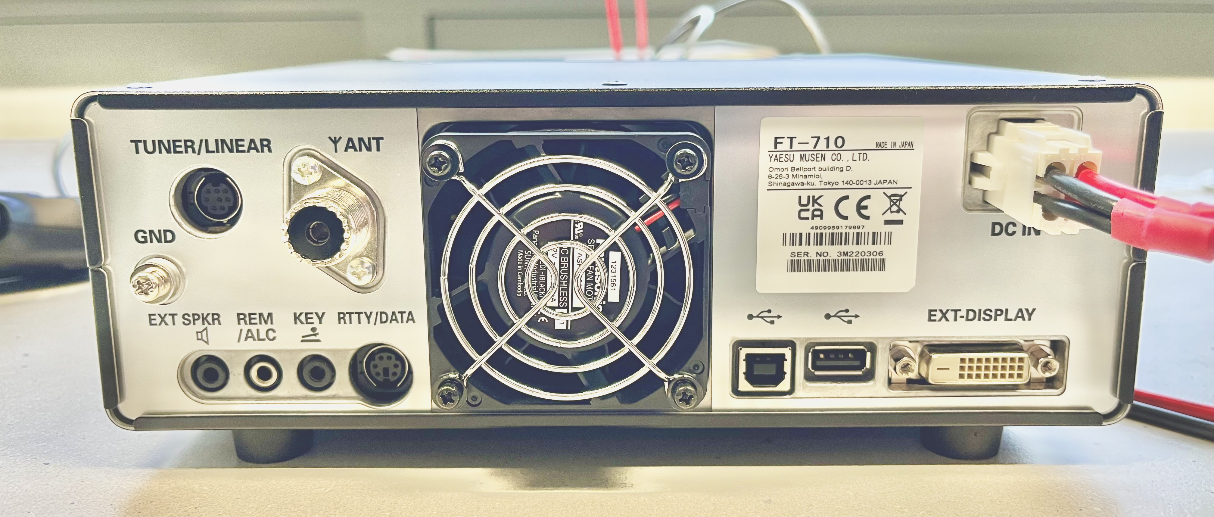 Dieser Alt-Text wurde noch nicht überprüft.
<ol>
<li>
<p>Kurze Zusammenfassung: Rückseite eines elektronischen Geräts (Modell FT‑710) mit vielen Anschlüssen, zentralem Lüftergitter und angeschlossenem DC‑Stromkabel.</p>
</li>
<li>
<p>Detaillierte Beschreibung: Das Gerät steht auf einem hellen Tisch; im Zentrum befindet sich ein großer, runder, mit vier Schrauben befestigter Lüfter mit Metall-Schutzgitter und sichtbarem „Brushless Fan“-Aufkleber. Links sind mehrere Buchsen und Beschriftungen: oben „TUNER/LINEAR“ an einer runden mehrpoligen Mini‑DIN‑Buchse, darunter „GND“ an einem Massebolzen mit Rändelmutter, daneben „Y ANT“ bei einer großen koaxialen SO‑239‑Buchse. In der unteren linken Reihe stehen nebeneinander Klinkenbuchsen und Mini‑DIN‑Buchsen, beschriftet mit „EXT SPKR“, „REM/ALC“, „KEY“ und „RTTY/DATA“. Rechts neben dem Lüfter klebt ein weißes Typenschild mit der Aufschrift „FT‑710“, Hersteller‑ und Zulassungszeichen sowie einer Seriennummer. Ganz rechts oben ist „DC IN“ mit einem weißen Steckverbinder zu sehen, in den rote und schwarze Stromkabel eingesteckt sind. Unten rechts ist der Bereich „EXT‑DISPLAY“ mit mehreren Schnittstellen: zwei USB‑Anschlüsse (links ein quadratischer Typ‑B, daneben ein rechteckiger Typ‑A), ein kleiner gewindeter Koax‑Anschluss und ein weißer DVI‑Anschluss mit zwei Befestigungsschrauben. Der Hintergrund ist unscharf und zeigt eine helle Umgebung."></p>
 <figcaption>Abbildung E-12.3.1: Funkgerät mit DATA-Port</figcaption>
</li>
</ol>
</figure>
<figure class=