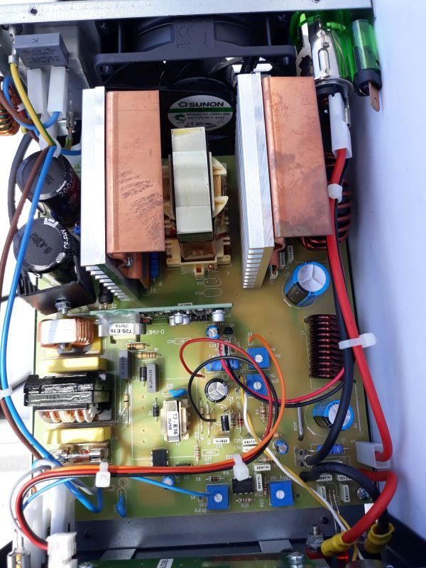 Dieser Alt-Text wurde noch nicht überprüft.
<ol>
<li>
<p>Kurze Zusammenfassung: Innenansicht eines geöffneten elektronischen Geräts mit bestückter Leiterplatte, großen Kühlkörpern, einem Lüfter, Spulen, Kondensatoren und mehreren farbigen Kabeln.</p>
</li>
<li>
<p>Detaillierte Beschreibung: Die Aufnahme zeigt eine gelbgrüne Leiterplatte in einem Metallgehäuse von oben. In der oberen Bildhälfte sitzt mittig ein schwarzer runder Lüfter mit sichtbarem „SUNON“-Aufkleber. Davor stehen zwei massive, kupferfarbene Blöcke auf silbernen Kühlrippen; zwischen ihnen ein beigefarbener, rechtwinkliger Transformator-Kern. Rechts oben befindet sich eine grüne Glasrohrsicherung in einer Halterung sowie dicke rote und schwarze Leitungen, mit weißen Kabelbindern am Rand fixiert. Über die Platine verteilt sind mehrere große Elektrolytkondensatoren in Blau und Schwarz, zahlreiche blaue Trimmpotentiometer, verschiedene Widerstände, Dioden und kleinere Halbleiter. Rechts unten liegt eine dunkelbraune Spule mit dicker lackierter Kupferwicklung; links sind weitere Wickelspulen und schwarze Bauteile mit Kühlblechen zu sehen. Am linken oberen Rand stehen weiße quaderförmige Komponenten, daneben verlaufen blaue und rote Leitungen. Die Leiterplatte trägt weiße Siebdruck-Beschriftungen und Markierungen; mehrere rote, gelbe, blaue und schwarze Einzeldrähte sind steckbar an den Rändern angeschlossen und kreuzen teils die Bildmitte."></p>
 <figcaption>Abbildung E-7.3.1: Innenansicht eines Schaltnetzteils für $13,8&nbsp;V$ und $35&nbsp;A$ mit einem kleinen Ferritkernübertrager zwischen den beiden Kühlkörpern, Gewicht $2&nbsp;k\gram$</figcaption>
</li>
</ol>
</figure>
</aside>
Das Schaltnetzteil besitzt mehrere positive Eigenschaften gegenüber einem linear geregelten Netzteil:
<ul>
<li>Hoher Wirkungsgrad auch bei kleinen Nennspannungen und wechselnden Lasten</li>
<li>Geringes Gewicht und Volumen, da durch die hohe Frequenz kleinere Transformatoren und sekundäre Siebkondensatoren verwendet werden</li>
<li>Gute Regelbarkeit</li>
<li>Geringeres Gewicht, kleinere Kühlkörper und dadurch weniger Platzbedarf im Vergleich zu einem linear geregelten Netzteil (siehe Abschnitt: Spannungsstabilisierung)</li>
</ul>
<section class=