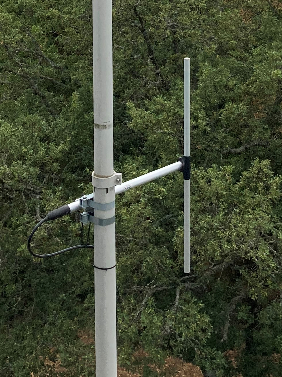 Dieser Alt-Text wurde noch nicht überprüft.
<ol>
<li>
<p>Kurzzusammenfassung: An einem hellen Mast ist eine kleine Antenne mit zwei parallelen vertikalen Stäben montiert, angeschlossen über ein schwarzes Koaxkabel; im Hintergrund dichter Baumbestand.</p>
</li>
<li>
<p>Detaillierte Beschreibung: Ein senkrechter, hellgrauer Mast verläuft links im Bild und zeigt leichte Gebrauchsspuren. Mit zwei Metallbandschellen ist daran ein kurzer, waagerechter Ausleger befestigt. Am freien Ende des Auslegers stehen zwei schlanke, helle Stäbe parallel nach oben; der hintere Stab ist länger, der vordere etwas kürzer und mit geringem Abstand davor. Am Übergang zwischen Mast und Ausleger sitzt eine kleine Halterung, in die ein schwarzes Koaxialkabel mit Stecker eingeführt ist; die Anschlusspunkte sind mit dunklem Band oder Schrumpfschlauch abgedichtet. Das Kabel führt in einer Schlaufe nach unten. Der Hintergrund besteht vollständig aus dichtem, grünem Laub von Bäumen; nahe dem unteren Bildrand ist etwas brauner Boden durch das Geäst zu erkennen."></p>
 <figcaption>Abbildung E-14.2.5: Vertikaler $\frac{\lambda}{2}$-Dipol</figcaption>
</li>
</ol>
</figure>
</aside>
---
<p>Einen Spezialfall einer Vertikalantenne stellt die gegen Erde (oder einer Fahrzeugkarosserie) erregte $5/8 \lambda$-Antenne dar (vgl. Abbildung <a class=