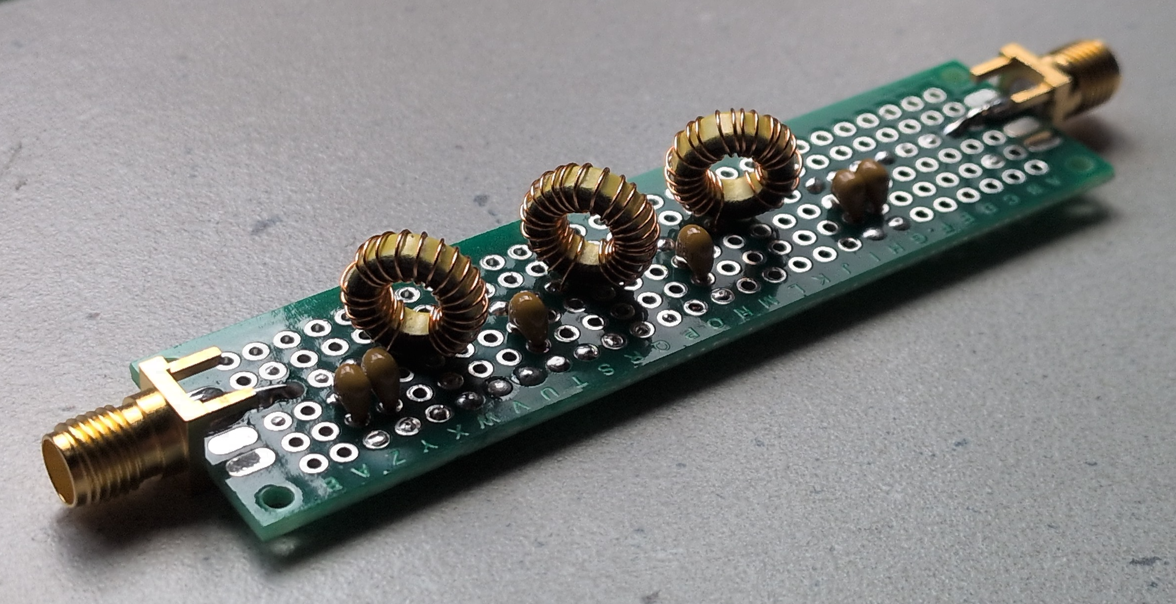 Dieser Alt-Text wurde noch nicht überprüft.
<ol>
<li>
<p>Zusammenfassung: Langer, schmaler Elektronikaufbau auf grünem Lochrasterboard mit drei ringförmigen Kupferspulen und mehreren kleinen braunen Bauteilen zwischen zwei goldfarbenen Koaxanschlüssen.</p>
</li>
<li>
<p>Detaillierte Beschreibung: Auf einem rechteckigen, grünen Lochrasterboard mit Reihen aus runden Lötaugen sind drei gleichartige, ringförmige Spulen aus lackiertem Kupferdraht auf gelblichen Kernen in einer Linie angeordnet. Dazwischen und daneben sitzen mehrere kleine, braun glänzende, tropfenförmige Bauteile (per Drahtanschluss eingelötet). An beiden Stirnseiten des Boards befinden sich goldfarbene, koaxiale Buchsen/Stecker mit verlöteten Mittelkontakten und seitlichen Lötfahnen zur Masse. Die Lötstellen sind sichtbar, teils mit dunklen Flussmittelspuren. Das Board liegt schräg auf einer grauen, leicht strukturierten Oberfläche; die Beleuchtung kommt von oben links und erzeugt weiche Schatten."></p>
 <figcaption>Abbildung NEA-11.1.10: Selbstgebautes Tiefpassfilter</figcaption>
</li>
</ol>
</figure>
Die oben genannten Filter lassen sich natürlich wunderbar für alle Frequenzbereiche selbst berechnen und bauen. In der Formelsammlung finden sich die benötigten Formeln, wobei es natürlich auch viele Bauvorschläge und Berechnungsprogramme gibt. Die benötigten Spulen lassen sich dazu oft leicht selbst herstellen. Hierfür reicht bei kleinen Spulenwerten ein kleiner Vorrat an $0,8&nbsp;mm$-Kupferlackdraht für stabile Luftspulen aus. Bei großen Spulenwerten z.B. für die Kurzwellenbereiche kommt man mit $0,2&nbsp;mm$-Kupferlackdraht und Kernmaterial mit entsprechenden $A_\text{L}$-Werten aus, um sich jederzeit die richtigen Werte selbst herstellen zu können. Die benötigten Größen, Wicklungen usw. bekommt man über Formelsammlung, Bauvorschläge oder Berechnungsprogramme ebenfalls meist leicht heraus.
</aside>
---
<p>Nun haben wir einfache RC- und LC-Glieder als Hoch- und Tiefpassfilter kennengelernt. Aus Kondensatoren und Spulen lassen sich jedoch noch weitere Filtertypen realisieren, die über reine Hoch- und Tiefpässe hinausgehen. Diese wollen wir uns nun im zweiten Teil genauer ansehen, nämlich die sogenannten <em>Schwingkreise</em>.</p>
<aside class=