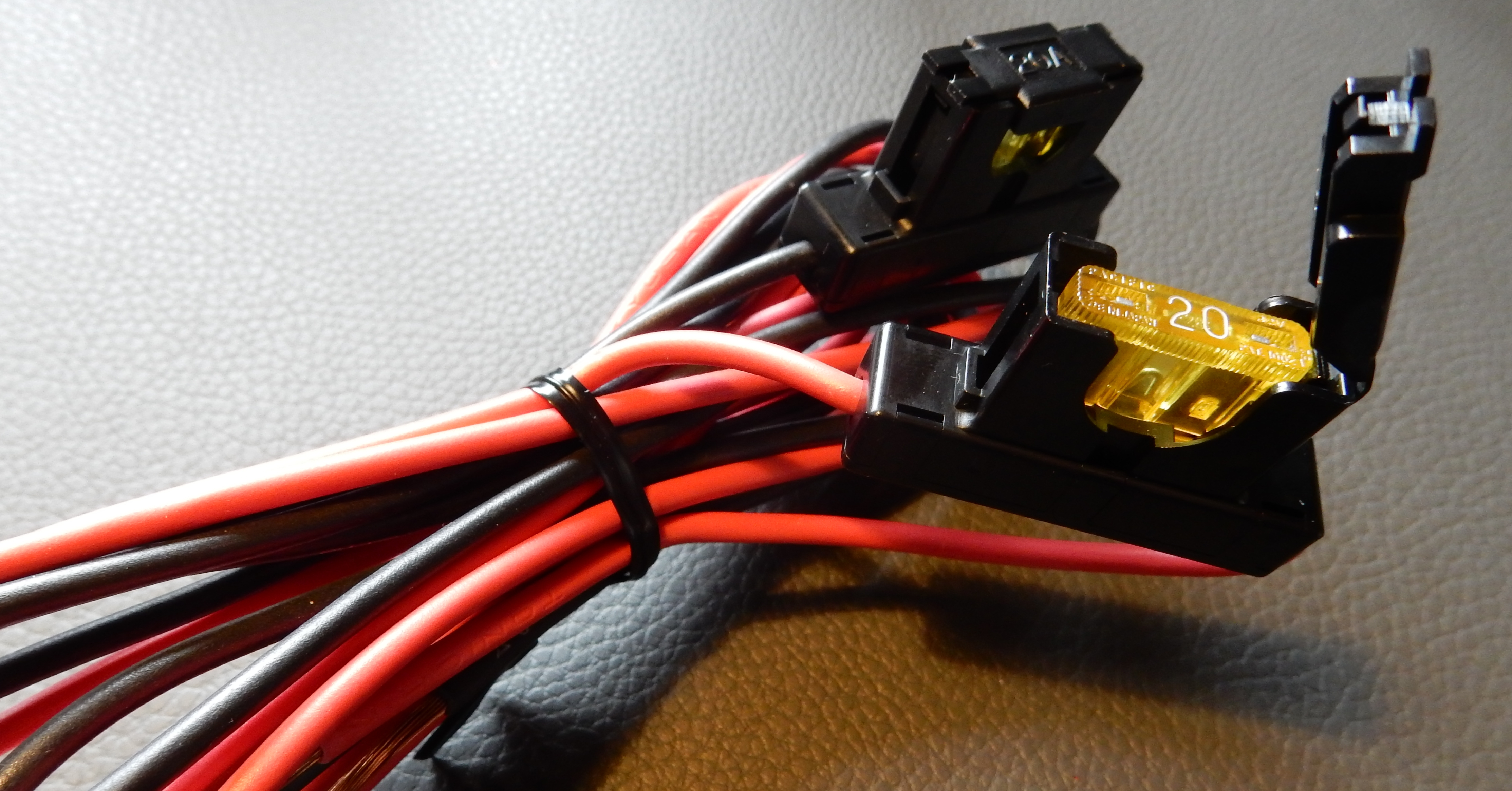 1) Kurzbeschreibung: Bündel roter und schwarzer Kabel mit zwei Inline-Sicherungen, von denen eine geöffnet ist und eine gelbe 20‑A‑Flachsicherung zeigt.
<ol start=